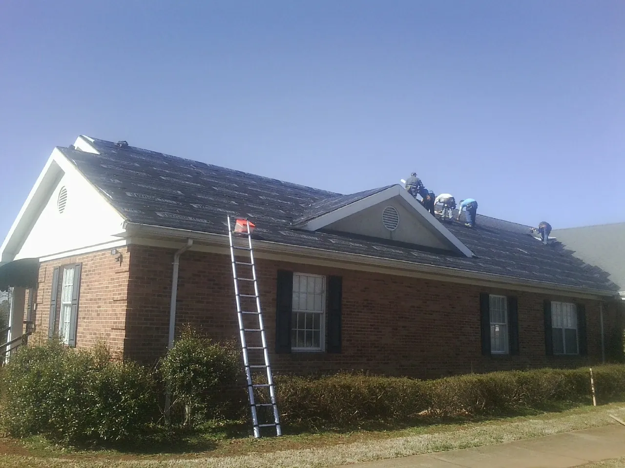 Crew tearing shingles off a brick ranch roof with a ladder set — Emergency Roof Repair work by our crew in Rhodhiss