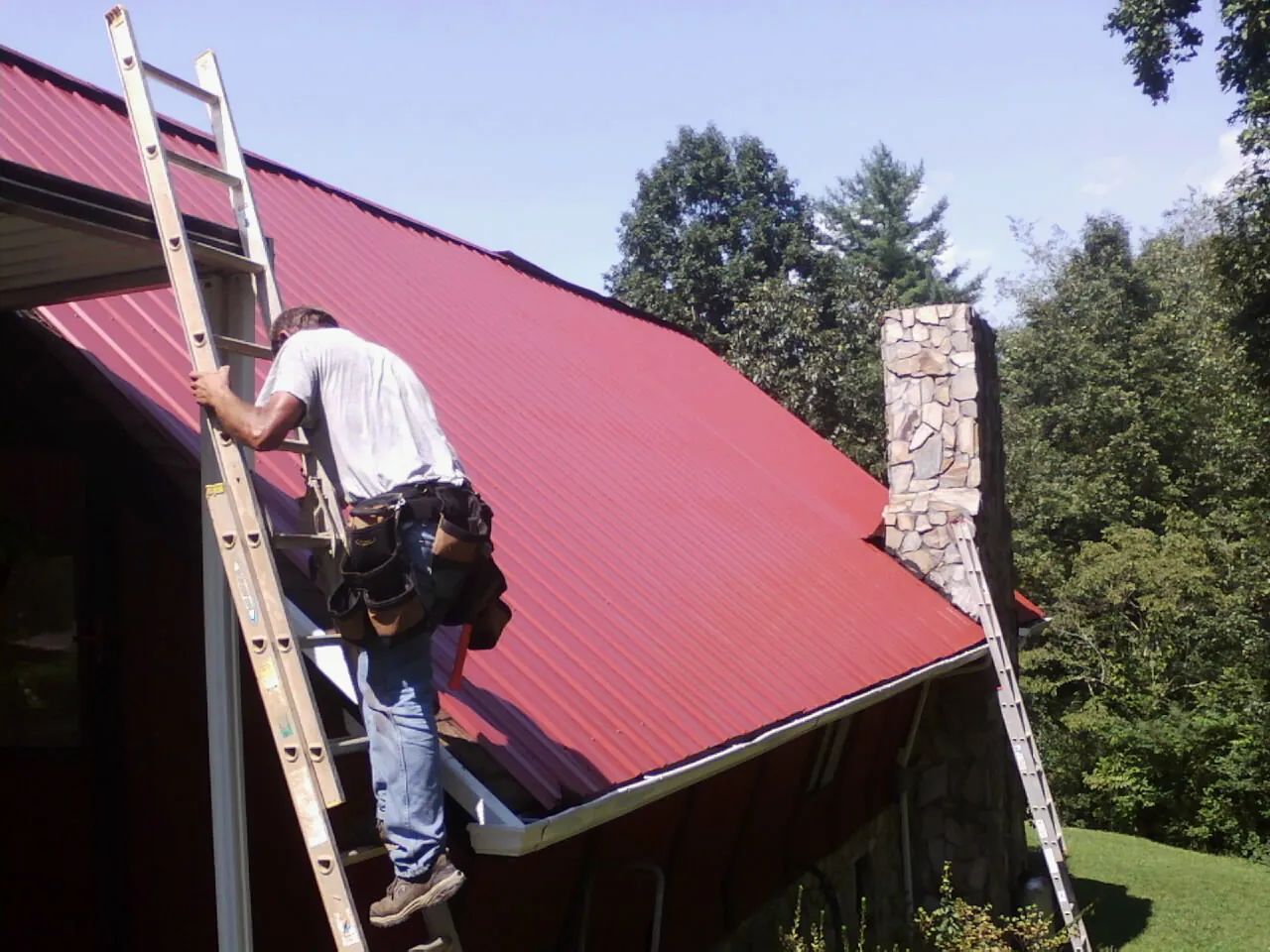 Roofer on a ladder beside a red metal roof and stone chimney — Soffit & Fascia Repair by Roofing Brothers in Rhodhiss